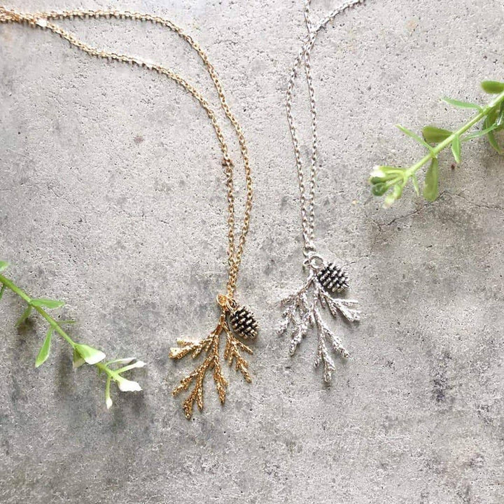 Two necklaces on a grey surface. One necklace has a gold-colored pine cone and branch pendant, and the other has a silver-colored pine cone and branch pendant. Both necklaces have delicate chains.