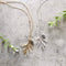 Two necklaces on a grey surface. One necklace has a gold-colored pine cone and branch pendant, and the other has a silver-colored pine cone and branch pendant. Both necklaces have delicate chains.