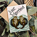 Card with 'Just Married' text and otter illustration, surrounded by greenery