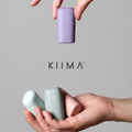 Two hands holding a purple and a light blue cylindrical product with 'KIIMA' branding on a gray background.