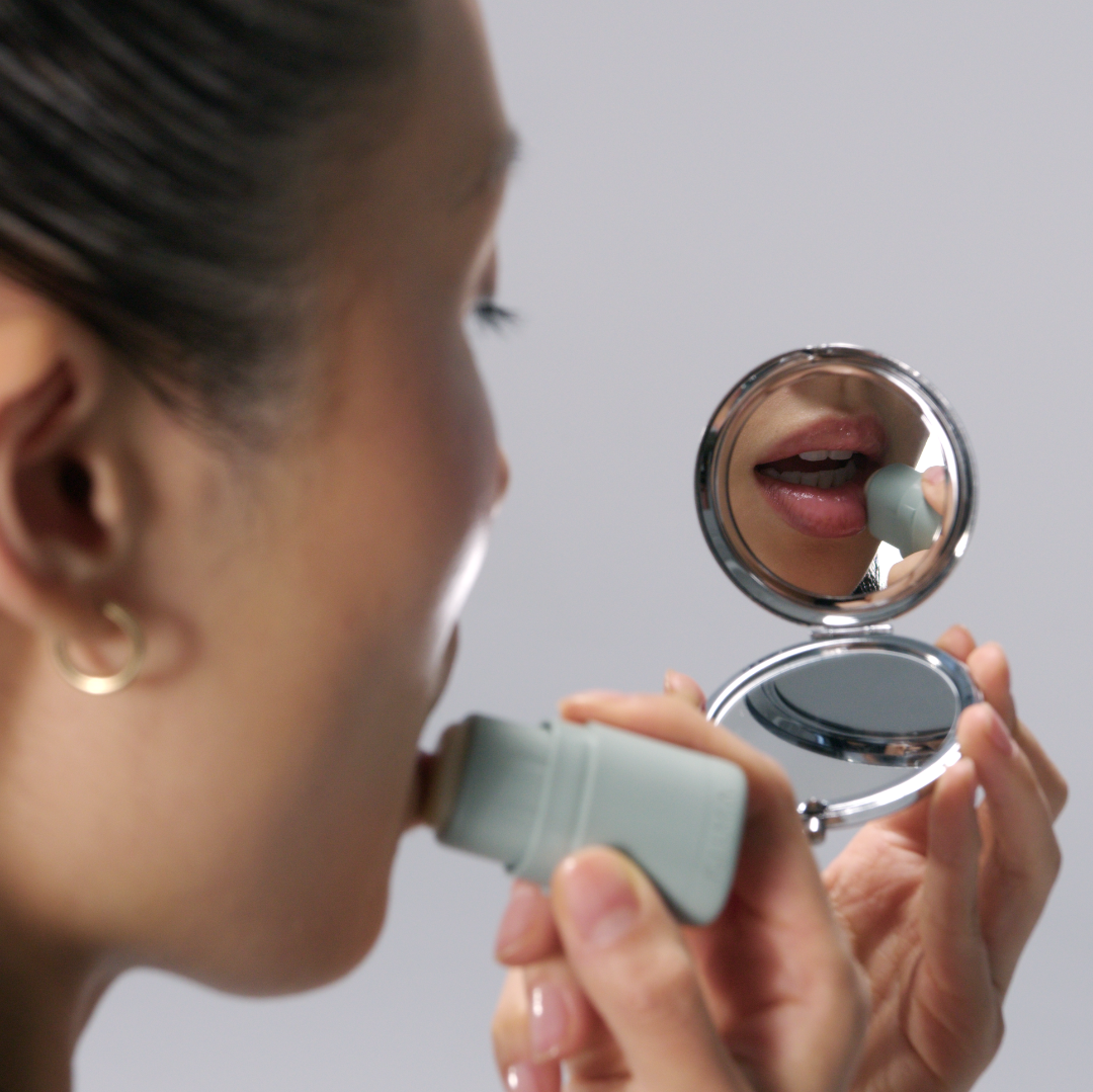 Woman applying makeup with a compact mirror on a plain background