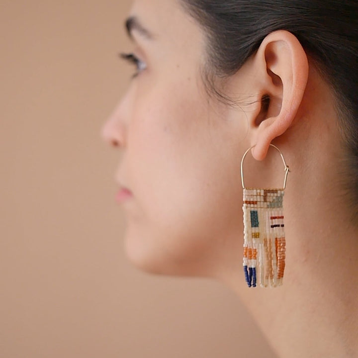 A pair of rectangular, tapestry-style earrings hand-stitched with small Japanese glass beads in an abstract, painterly pattern featuring shades of cream, red, burgundy, orange, yellow, teal, and blue. They hang from delicate, half-circle 14 KT gold filled wire hoops.