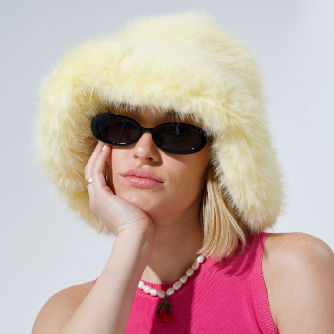 Woman wearing a fluffy yellow hat and sunglasses with a pink top against a light background