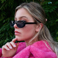 Woman wearing black sunglasses and a pink coat against a green leafy background