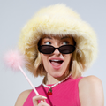 Person wearing a fluffy yellow hood, sunglasses, and a pink dress with a red bow, holding a pink wand against a light gray background.