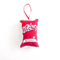 A close-up, high-angle flat lay shot on a plain white background features a soft, plush Christmas ornament shaped like a bag of ketchup chips. The ornament is bright red with white detailing, and the graphic features the word "Ketchup" in a stylized font, along with an image of a bottle of ketchup and a tomato. The bottom of the design is labeled "POTATO CHIPS CROUSTILLES". A red ribbon loop is attached to the top for hanging.