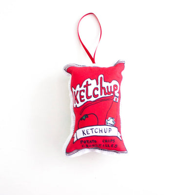 A close-up, high-angle flat lay shot on a plain white background features a soft, plush Christmas ornament shaped like a bag of ketchup chips. The ornament is bright red with white detailing, and the graphic features the word 
