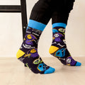 A person wearing black pants and bright blue crew socks with a spooky cartoon print, sitting on a black stool in a studio setting.