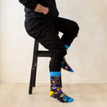 A man wearing a black hoodie, black pants, and colorful patterned crew socks sits on a black stool against a plain beige wall.