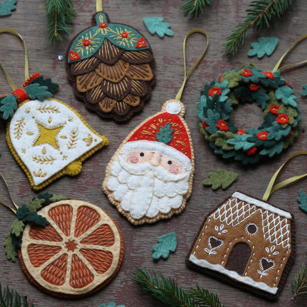 Six embroidered Christmas felt ornaments arranged on a rustic wooden surface with pine branches and small green leaves. The ornaments include a white bell with a gold star, a brown pinecone, a Santa Claus face, a green wreath with red berries, a sliced orange, and a gingerbread house.