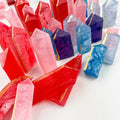 A collection of crystal-shaped candies in various colors, including pink, purple, blue, and red. Some candies have gold accents. The candies are arranged in a pile on a white surface.