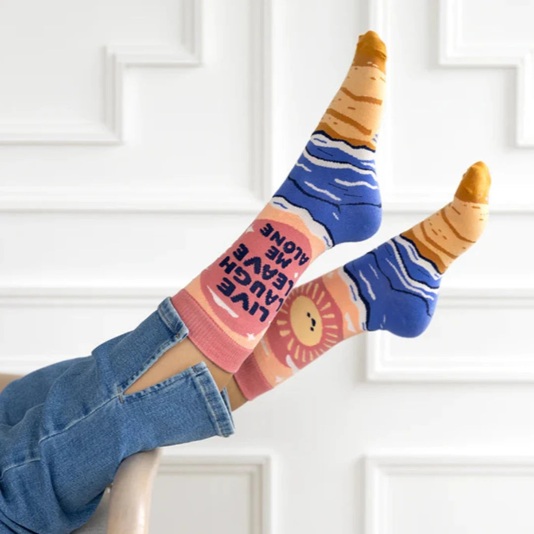 A pair of pink socks with a beach-themed design. The socks feature a beach scene, waves, and the text "LIVE LAUGH LEAVE ME ALONE."