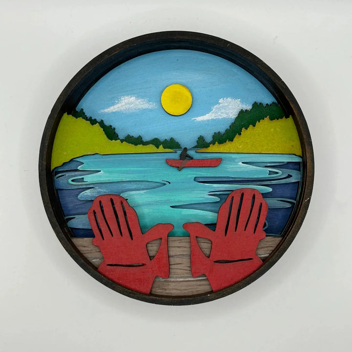 A Paint By Layers Craft Kit featuring a circular shadow box with a pre-designed landscape scene. The scene depicts a lake at sunset, with a person in a canoe and trees on the shore. The kit includes layers of wood, paint, and instructions. The title of the kit is 