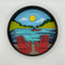 A Paint By Layers Craft Kit featuring a circular shadow box with a pre-designed landscape scene. The scene depicts a lake at sunset, with a person in a canoe and trees on the shore. The kit includes layers of wood, paint, and instructions. The title of the kit is 