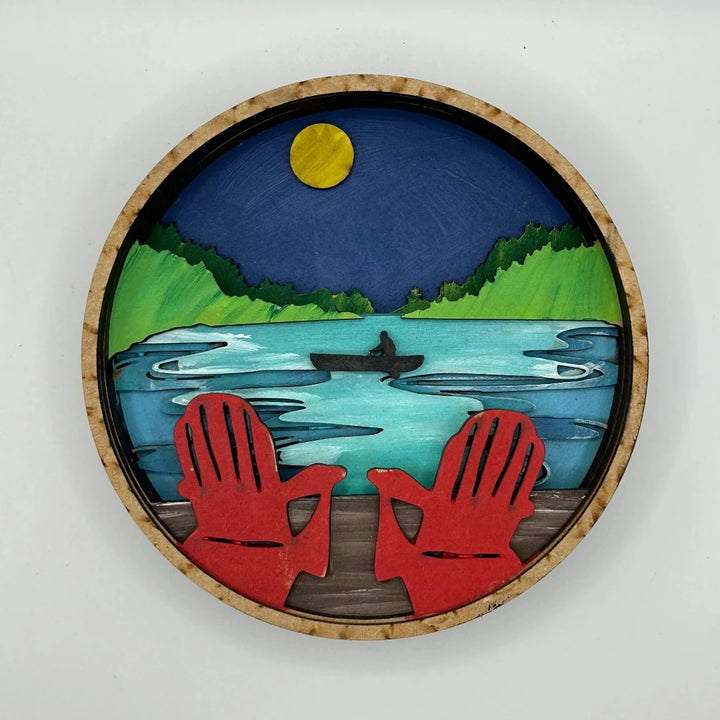 A Paint By Layers Craft Kit featuring a circular shadow box with a pre-designed landscape scene. The scene depicts a lake at sunset, with a person in a canoe and trees on the shore. The kit includes layers of wood, paint, and instructions. The title of the kit is 