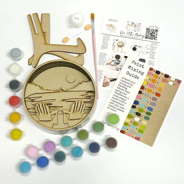 A Paint By Layers Craft Kit featuring a circular shadow box with a pre-designed landscape scene. The scene depicts a lake at sunset, with a person in a canoe and trees on the shore. The kit includes layers of wood, paint, and instructions. The title of the kit is 