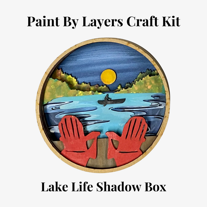 A Paint By Layers Craft Kit featuring a circular shadow box with a pre-designed landscape scene. The scene depicts a lake at sunset, with a person in a canoe and trees on the shore. The kit includes layers of wood, paint, and instructions. The title of the kit is 