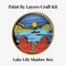 A Paint By Layers Craft Kit featuring a circular shadow box with a pre-designed landscape scene. The scene depicts a lake at sunset, with a person in a canoe and trees on the shore. The kit includes layers of wood, paint, and instructions. The title of the kit is 