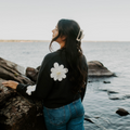 Person wearing a black jacket with a white flower design by a body of water.