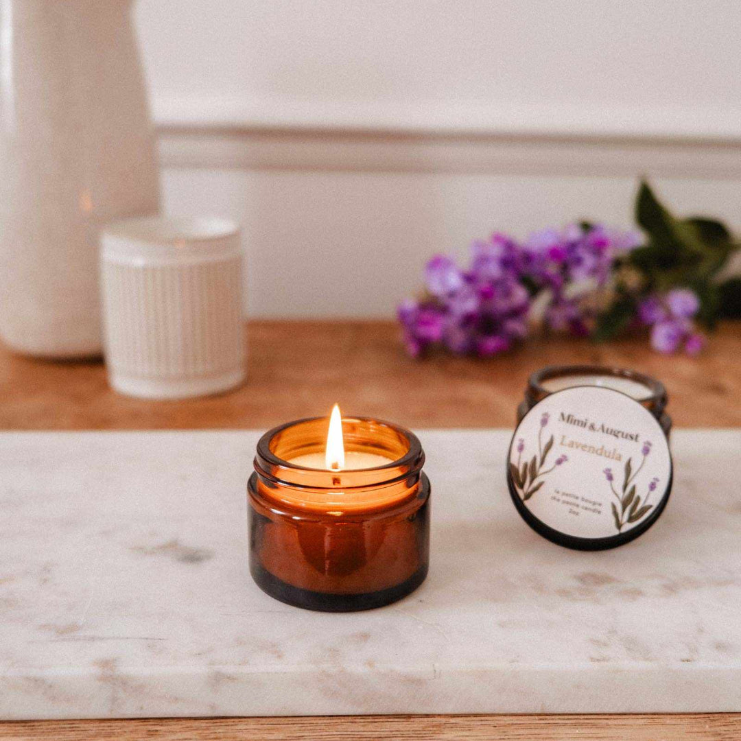 Lit amber glass candle on a marble surface with a white vase and purple flowers in the background.