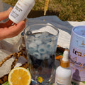 Person holding a bottle of 'Teas' product over a glass with ice and lemon, outdoor setting.
