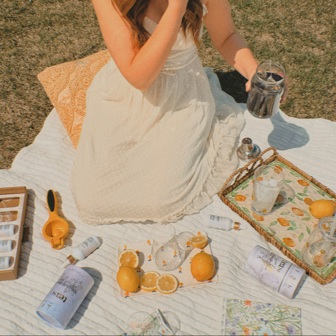Woman in a white dress sitting on a picnic blanket with lemons, bottles, and a tray.