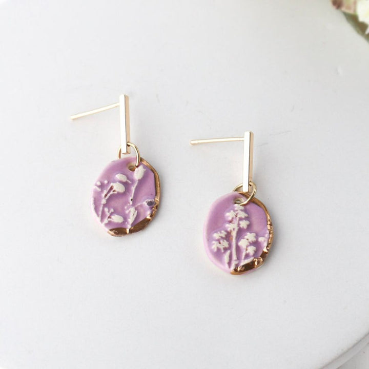 A pair of tiny, oval dangle earrings with a soft lavender glaze and a delicate white imprinted floral design, accented with a touch of luxurious 22K gold detailing around the edges. They hang from simple gold-toned 14K plated posts.