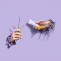 Two hands holding a tube of lip balm with lavender flowers on a purple background