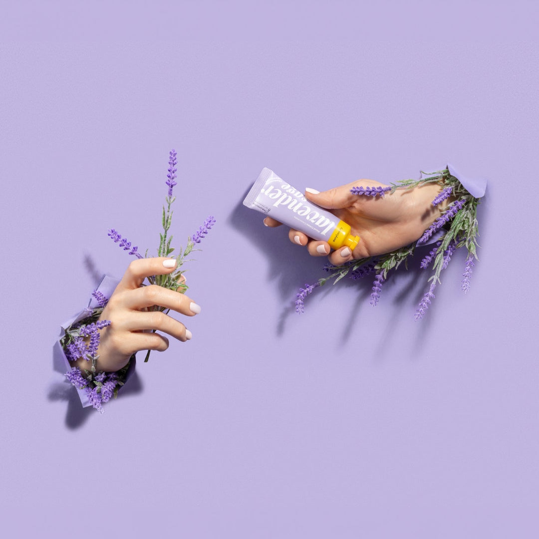 Two hands holding a tube of lip balm with lavender flowers on a purple background