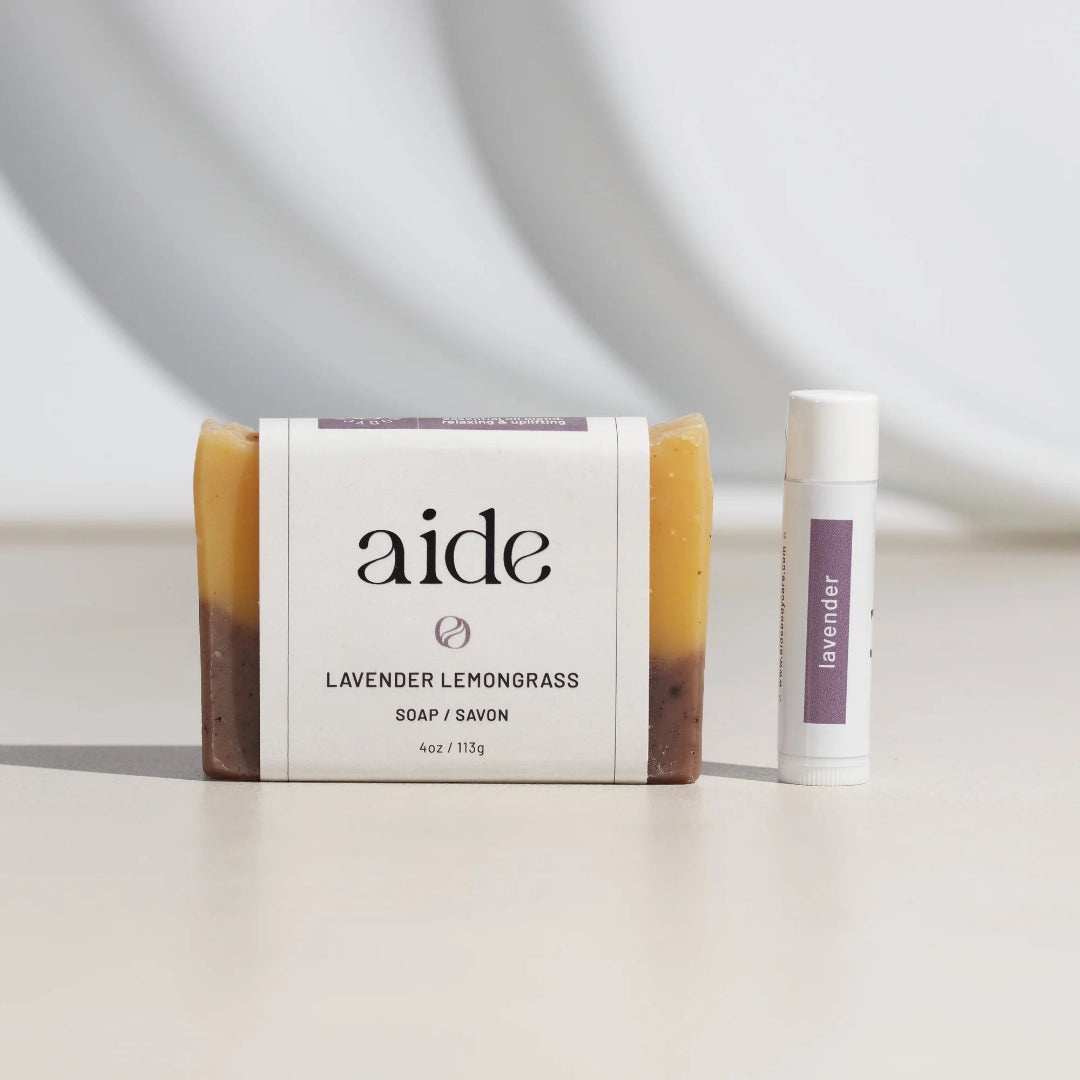 Bar of lavender lemongrass soap and a stick of lavender lip balm on a light surface with a neutral background.