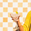 Hand holding a cocktail glass with a lime wedge against a yellow and white checkered background
