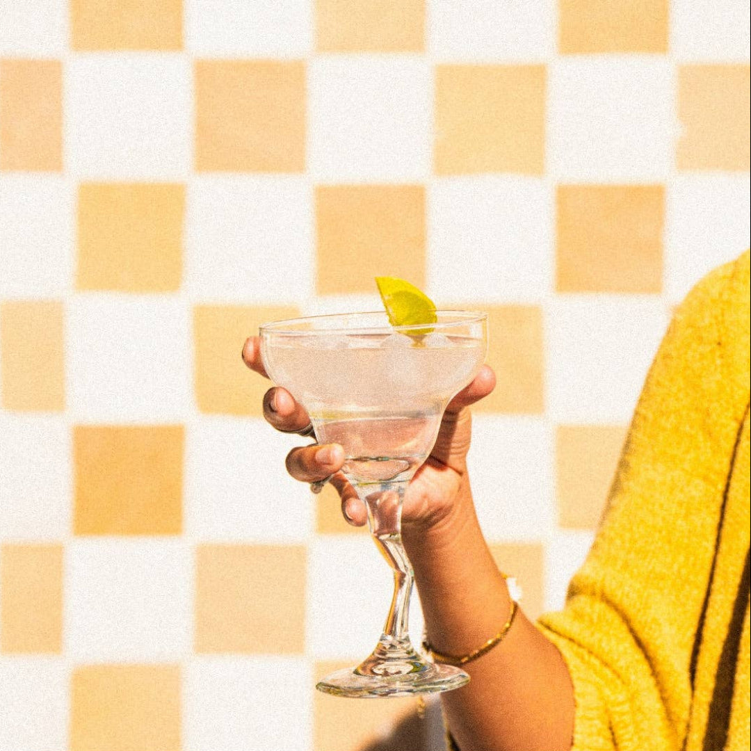 Hand holding a cocktail glass with a lime wedge against a yellow and white checkered background