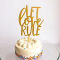 Small round cake with colorful sprinkles and a gold 'Let Love Rule' topper on a white background