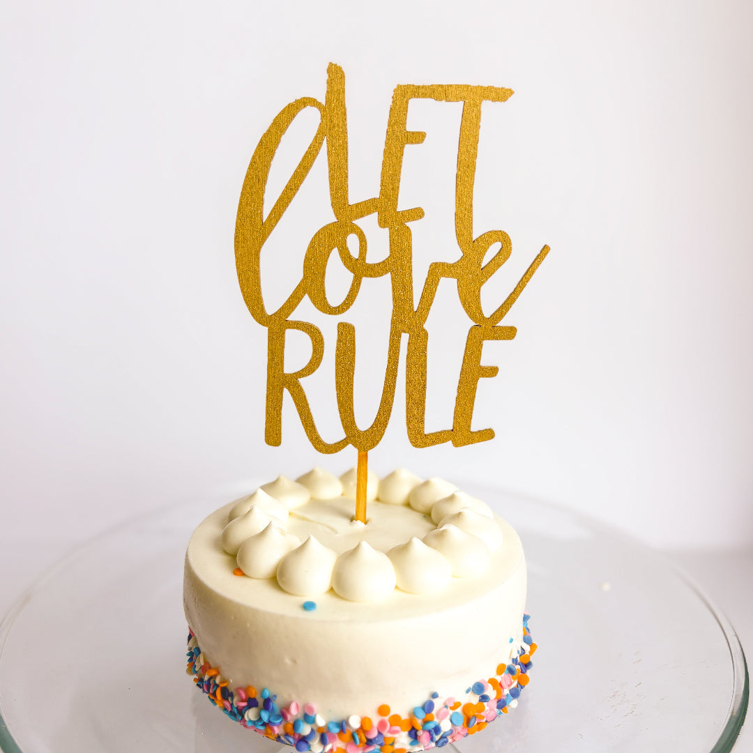 Small round cake with colorful sprinkles and a gold 'Let Love Rule' topper on a white background