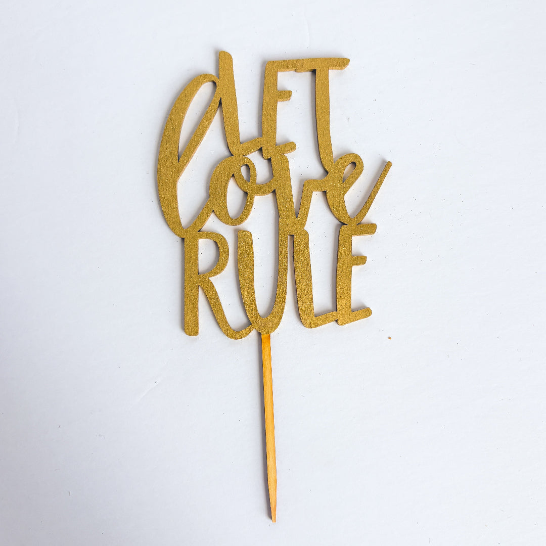 Gold cake topper with 'Let Love Rule' text on a white background