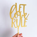 Gold glittery 'Let Love Rule' cake topper held against a white background