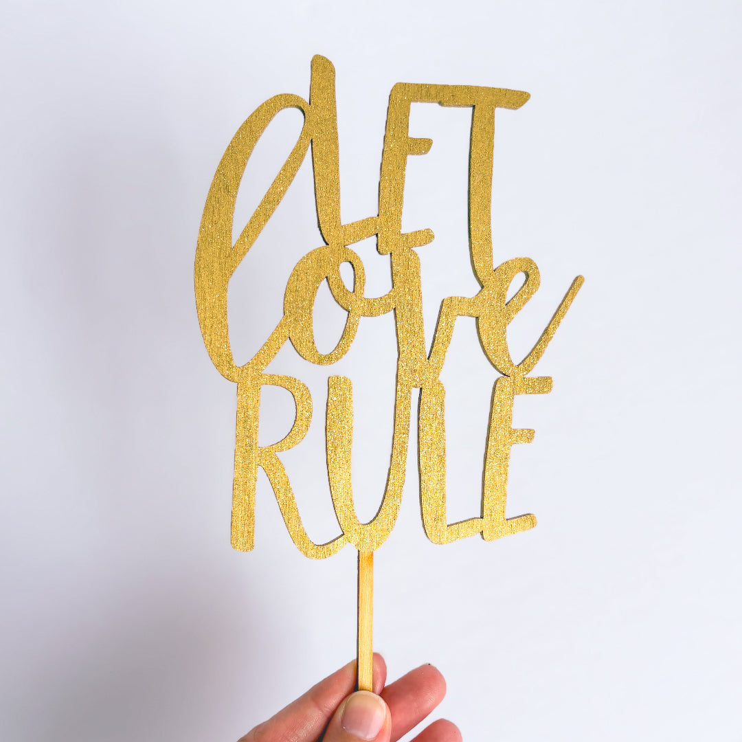 Gold glittery 'Let Love Rule' cake topper held against a white background