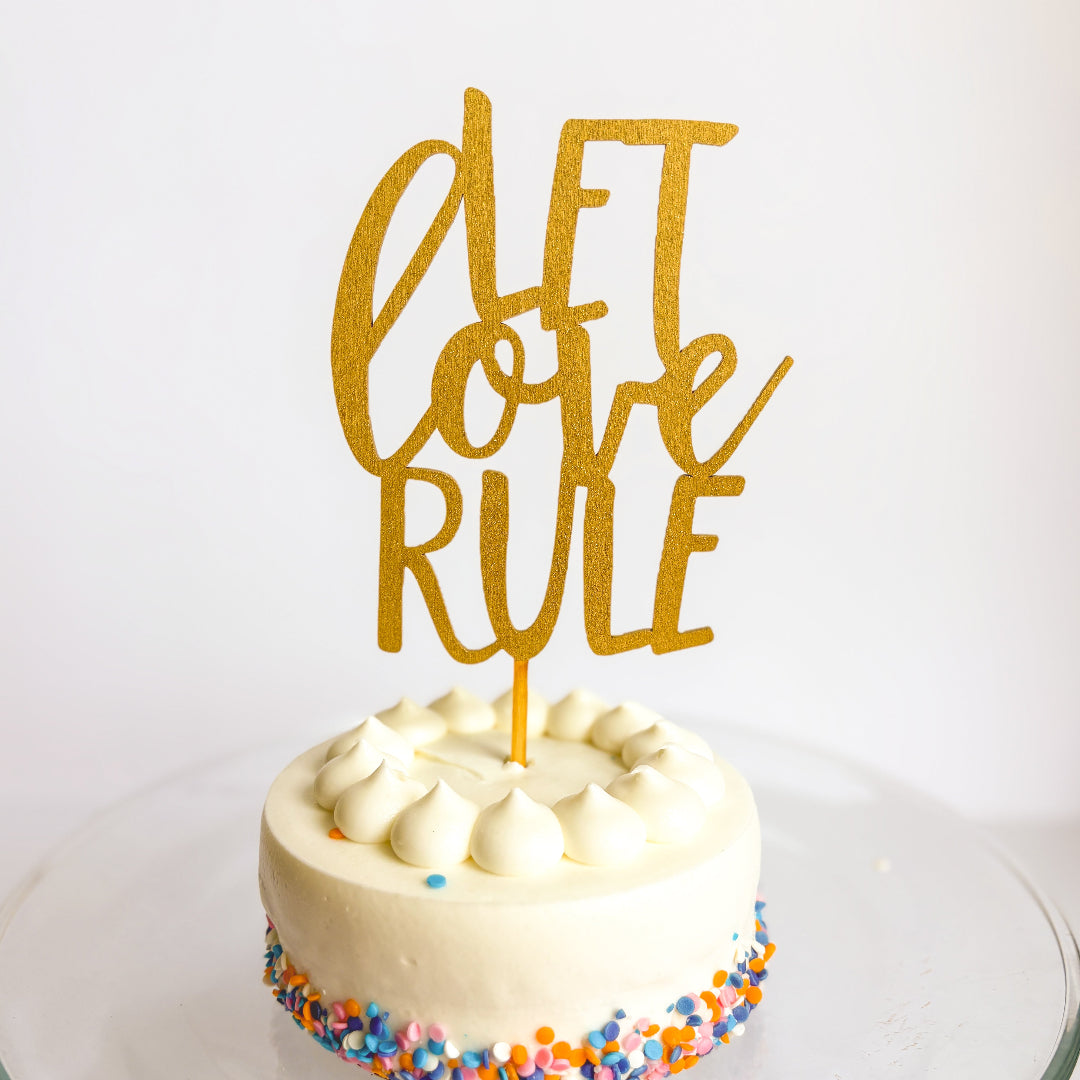 White cake with colorful sprinkles and a gold 'Let Love Rule' topper on a white background
