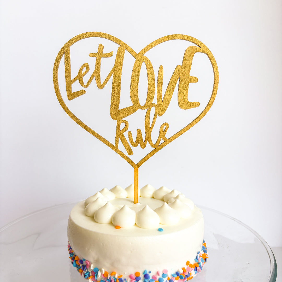 Small white cake with colorful sprinkles and a gold 'Let Love Rule' heart-shaped topper on a white background.
