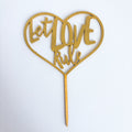Heart-shaped gold cake topper with 'Let Love Rule' text on a white background