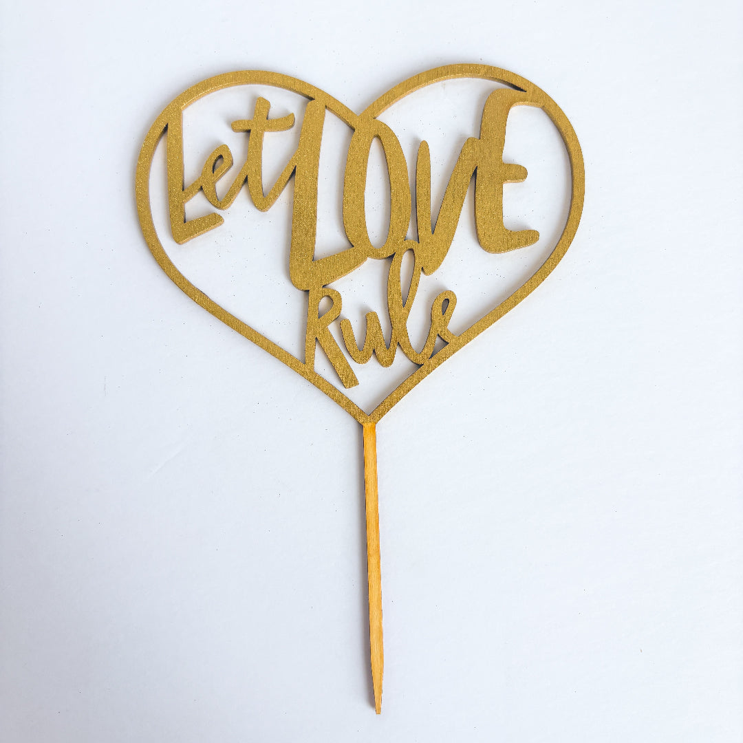 Heart-shaped gold cake topper with 'Let Love Rule' text on a white background