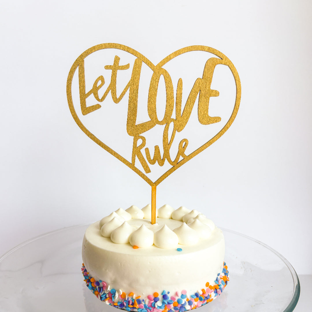 Gold 'Let Love Rule' heart-shaped cake topper on a white cake with colorful sprinkles.