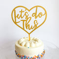 Gold heart-shaped cake topper with 'Let's do this' text on a white cake with colorful sprinkles.