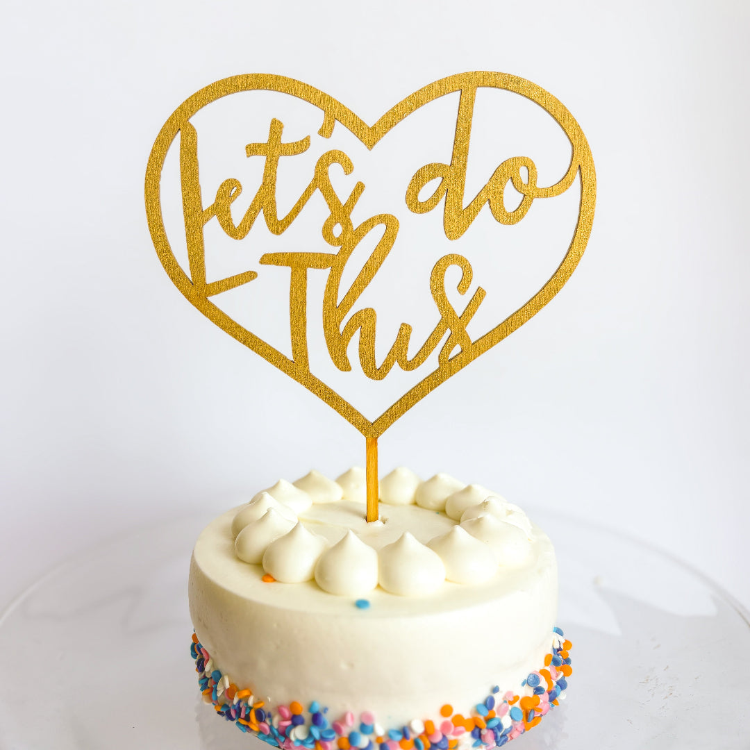 Gold heart-shaped cake topper with 'Let's do this' text on a white cake with colorful sprinkles.