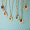 A collection of gold necklaces with colored gemstone pendants displayed on a light blue surface. Each pendant has a word engraved on it, such as 
