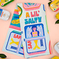 Colorful product packaging with 'A Lil Salty' text and illustrations on a light pink background.