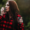 Woman in a red and black plaid shirt holding two small devices against a blurred natural background
