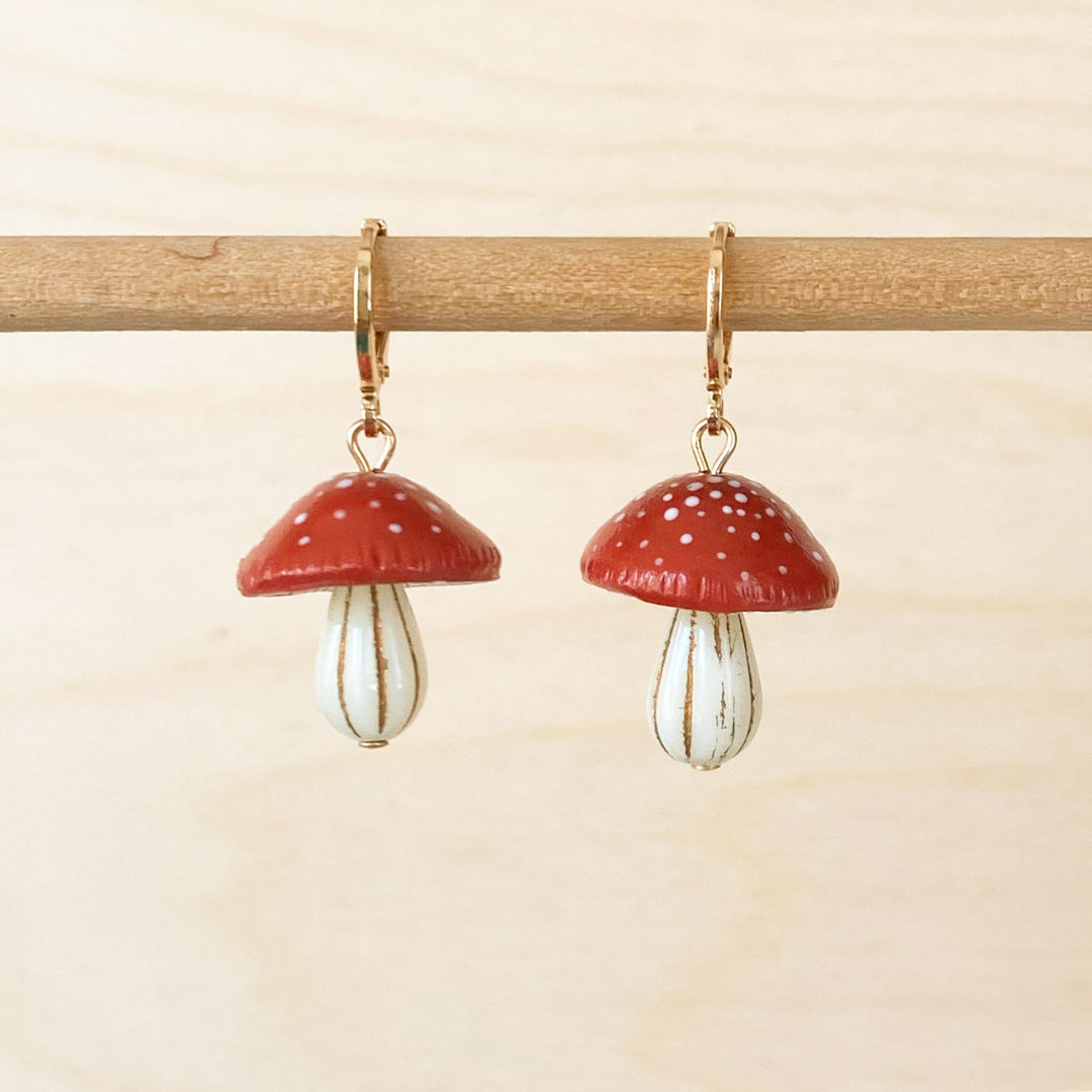 Mushroom-shaped earrings hanging on a wooden stick against a beige background
