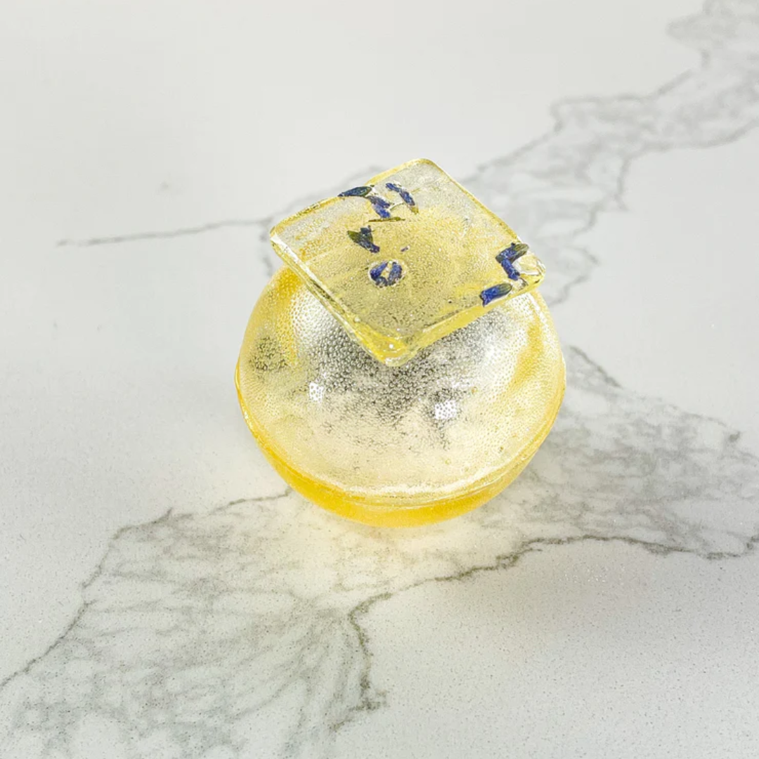 A yellow, spherical tea bomb topped with a square piece of edible glitter. The tea bomb is resting on a white marble surface.