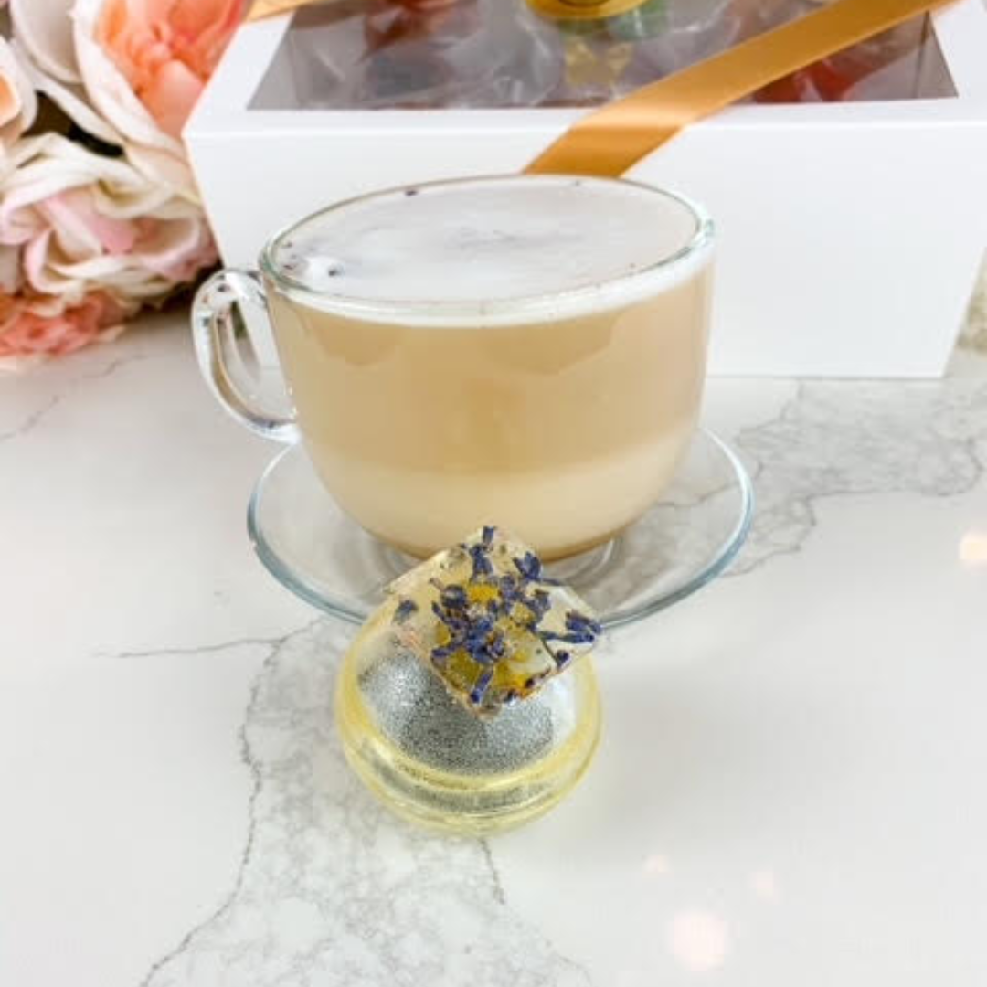 A yellow, spherical tea bomb topped with a square piece of edible glitter. The tea bomb is resting on a white marble surface.